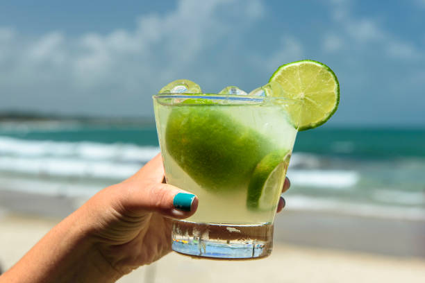 カイピリーニャを飲む。背景がぼやけたブラジルのビーチでカイピリーニャの飲み物を持っている手。 - drink alcohol summer celebration ストックフォトと画像