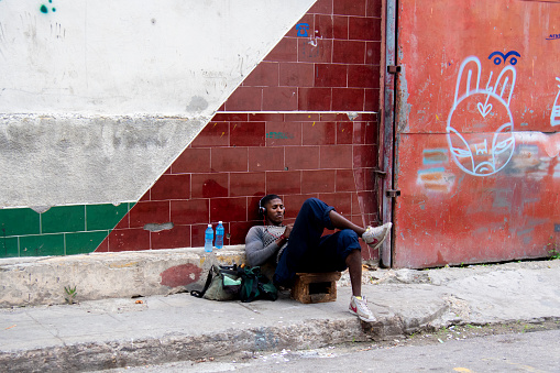 Young Cuban man listens to music on the headphone of his cellphone while relaxing lying down next to a mural drawing on the wall on a street in Havana, Cuba.