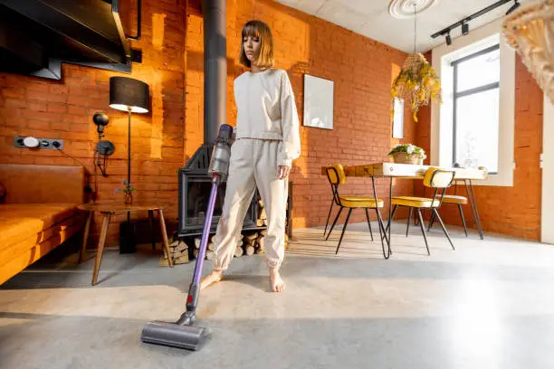 Woman cleaning concrete floor with cordless handheld vacuum cleaner in living room at home. Housewife doing housework