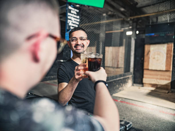 Coupe of young gay man at the axe throwing game room, taking a break from competition. . They are  dressed in casual clothing, wearing eyeglasses. Interior of warehouse like  space during the day.