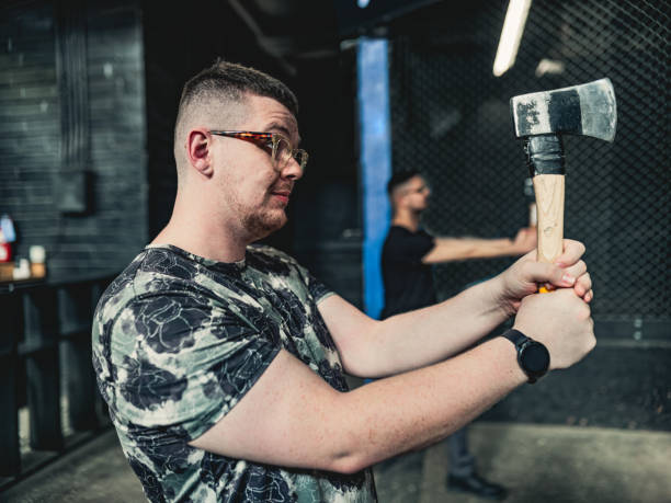 Young gay man throwing axe at the game range Young Caucasian gay man throwing axe at the game range. He is dressed in casual clothing, wearing eyeglasses. Interior of warehouse like  space during the day. axe throwing stock pictures, royalty-free photos & images