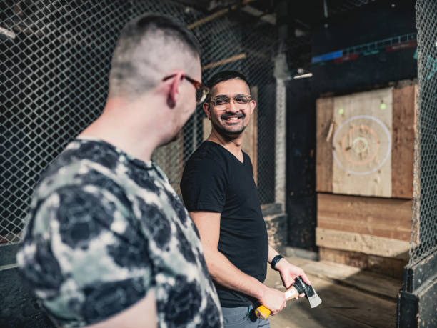 Young gay men at the axe throwing game room Coupe of young gay man at the axe throwing game room, competing with each other. They are  dressed in casual clothing, wearing eyeglasses. Interior of warehouse like  space during the day. axe throwing stock pictures, royalty-free photos & images