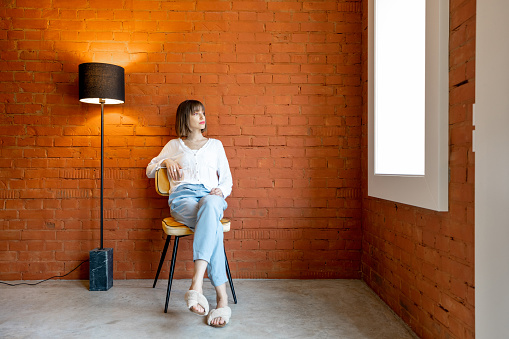 Full length portrait of a young woman sits on chair near floor lamp on brick wall background at home