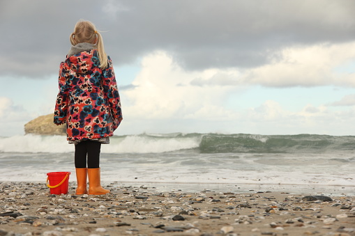 Young girl standing next to red bucket on sand and pebble beach looking out to breaking waves on sea