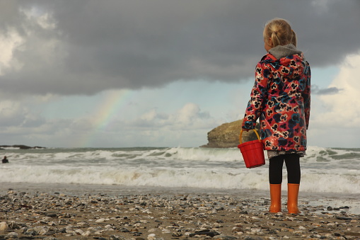 Young girl holding red bucket looking out to sea with breaking waves and rainbow in sky