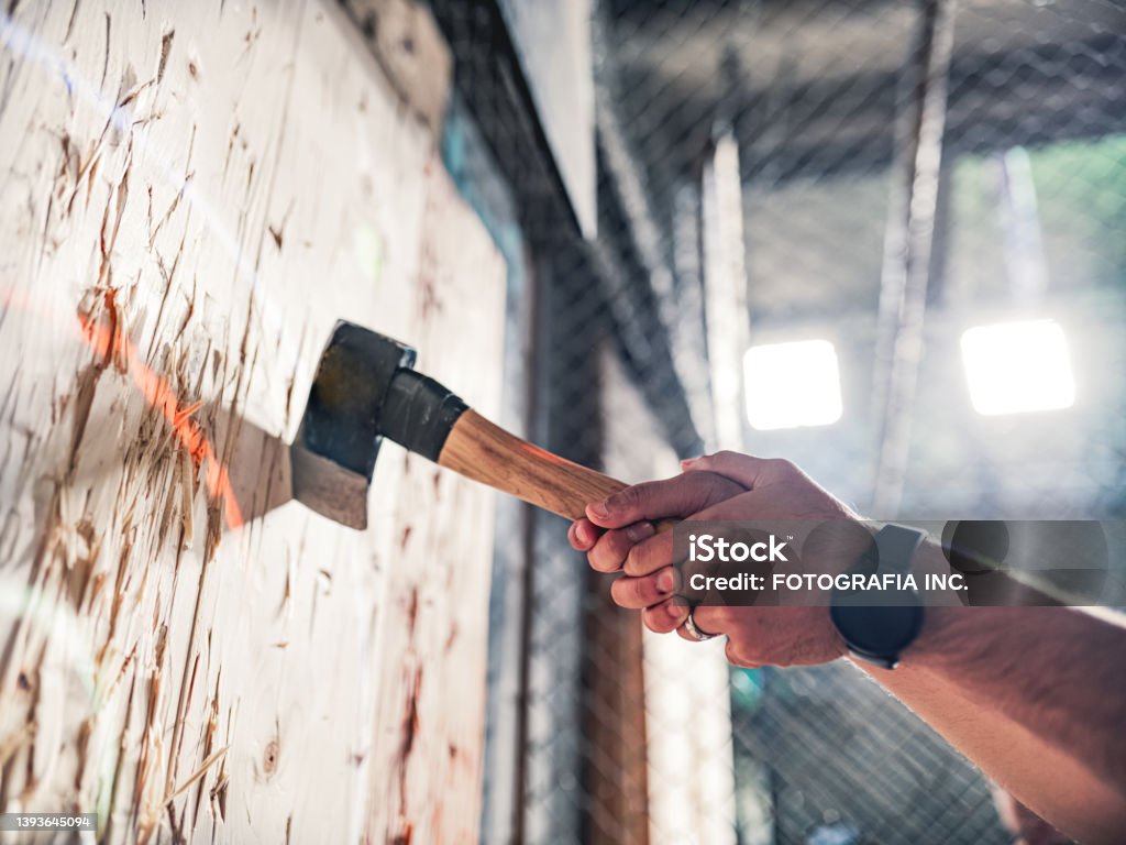 Young gay man throwing axe at the game range Hand of a Young Middle Easter gay man throwing axe at the game range. He is dressed in casual clothing. Interior of warehouse like  space during the day. Axe Throwing Stock Photo