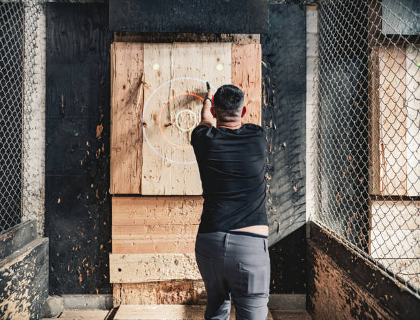 Young gay man throwing axe at the game range Young Middle Easter gay man throwing axe at the game range. He is dressed in casual clothing, wearing eyeglasses. Interior of warehouse like  space during the day. axe throwing stock pictures, royalty-free photos & images