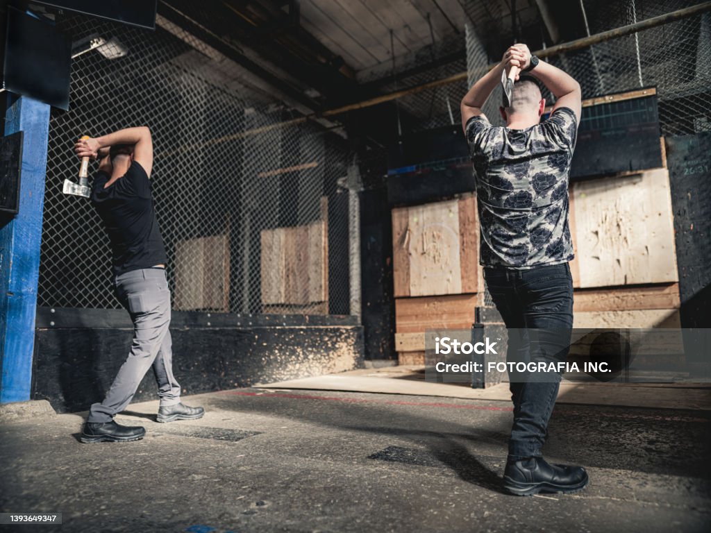 Young gay men at the axe throwing game room Coupe of young gay man at the axe throwing game room, competing with each other. They are  dressed in casual clothing, wearing eyeglasses. Interior of warehouse like  space during the day. Axe Stock Photo