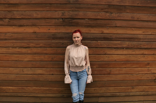 A Caucasian woman standing in front of a weathered brown wood wall. She is wearing a pink sleeveless top, torn blue jeans and black boots.