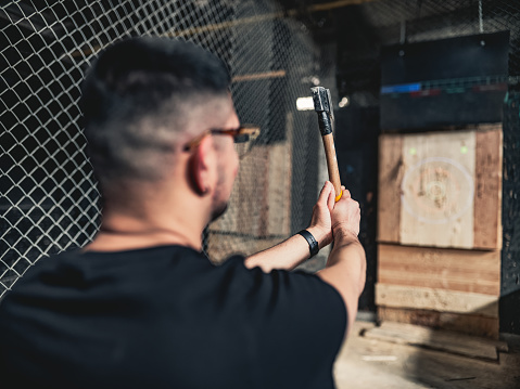 Young Middle Easter gay man throwing axe at the game range. He is dressed in casual clothing, wearing eyeglasses. Interior of warehouse like  space during the day.