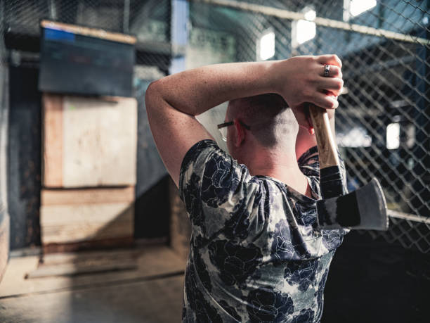 Young Caucasian gay man throwing axe at the game range. He is dressed in casual clothing, wearing eyeglasses. Interior of warehouse like  space during the day.
