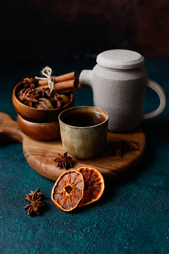 Istanbul, Turkey-April 11, 2022: Black tea in a small khaki green and rust effect ceramic cup without handle on a round wooden cutting board on a dark turquoise green rough floor. The background is black-brown. Behind the cup is a small gray stone teapot and walnuts and cinnamon sticks on wooden plates. There are dried orange slices and star anise in the front of the cup. Shot with Canon EOS R5.