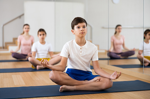 Concentrated teen boy sitting on mat in fitness center, making yoga meditation in lotus pose while exercising with family