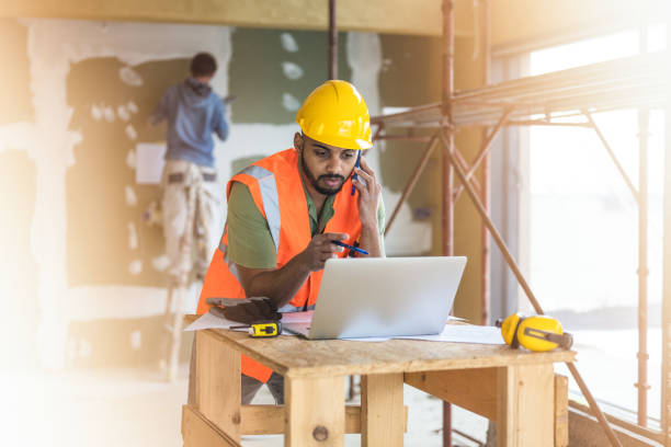 Man wearing hardhat and reflective vest looking at laptop and having call stock photo