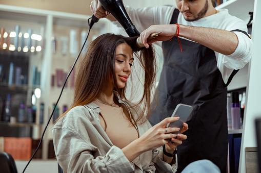 Beautiful woman with long hair using smart phone at beauty salon. Hair styling concept