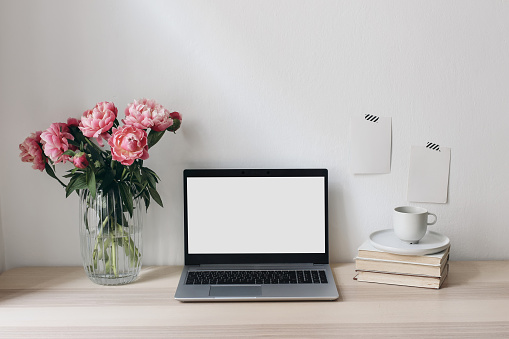 Home office still life scene. Notebook, computer and memo cards mock-ups. Vase with pink peonies flowers, books and cup of coffee. Wooden table, elegant feminine working space. Living room interior.