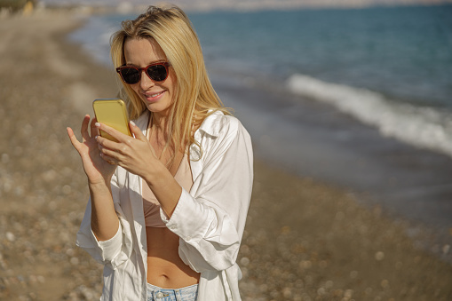 Blonde lady in sunglasses using mobile app while spending time on a beach. Vacation and technology