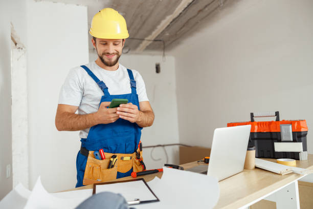 istock Joyful handyman in workwear and hardhat smiling while using smartphone, standing indoors during renovation work 1393677004