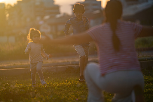 Photo of mother sitting arms outstretched and waiting to hug daughter and son running toward her in public park. Shot during sunset.