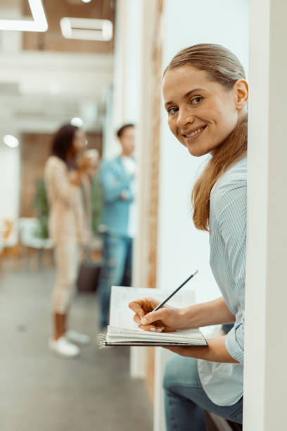 istock Smiling young female sitting on the windowsill and holding a notebook 1393686834