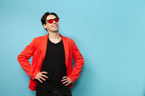 Studio portrait of a 20 year old man with brown hair in a pink jacket with pink sunglasses on a blue background