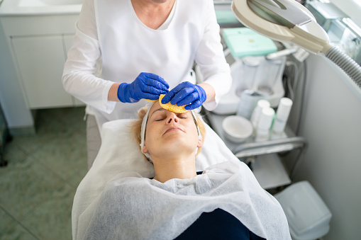 A beautician is using sponges to cleanse the facial treatment from her client, as the customer relaxes with her eyes closed