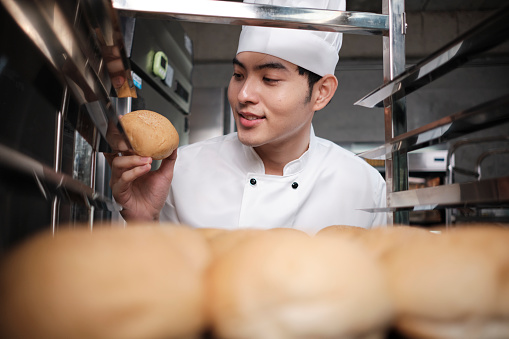 Young Asian male chef in white cook uniform and hat showing tray of fresh tasty bread with a smile, looking at his bun, happy with his baked food products, professional job at stainless steel kitchen.