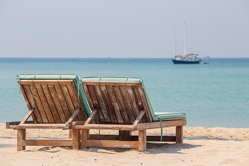 Two wooden deckchairs on a tropical sand beach overlooking the sea water and yacht. Thailand. Close up. Holiday and summer concept
