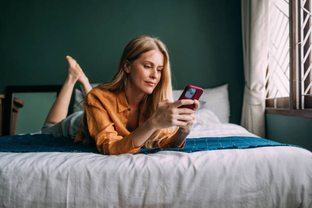 una bella donna bionda vestita casualmente guardando il suo smartphone rosso mentre si rilassa a casa dopo una lunga giornata - red bed foto e immagini stock