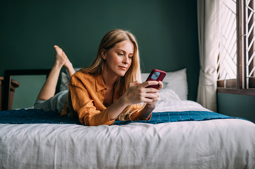 A charming Caucasian woman smiling while lying on her cozy bed and chatting on her mobile phone.