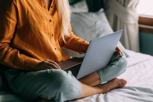 An Unrecognizable Blonde Female Working From Home On Her Laptop While Sitting On The Bed