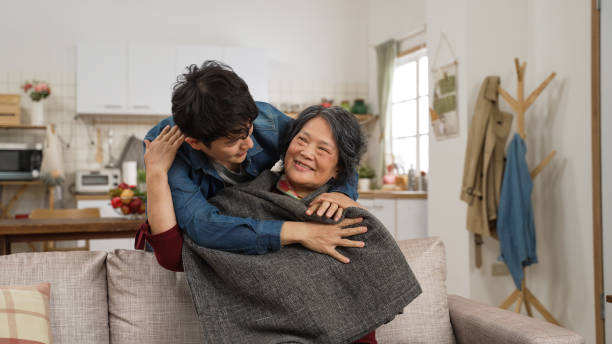 caring asian adult son coming over wrapping his mother up with a blanket as she is enjoying a fun tv show in the living room at home caring asian adult son coming over wrapping his mother up with a blanket as she is enjoying a fun tv show in the living room at home warm hug by chinese mom stock pictures, royalty-free photos & images