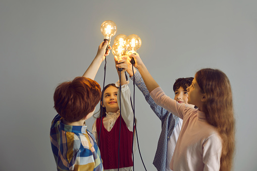 Team of children join glowing lightbulbs. Studio shot of intelligent little kids holding light bulbs. Sharing clever idea, support, collaboration, partnership, making difference, connection, teamwork