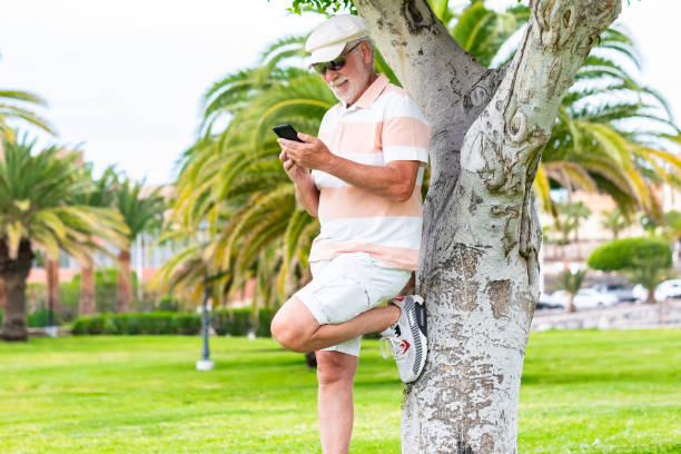 istock Beautiful bearded senior man standing under a tree in public park wearing hat and sunglasses using mobile phone. Smiling elderly man holding telephone using app and social media 1393755000