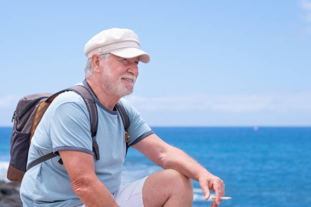 istock Attractive senior man with beard and hat sitting outdoors at sea looking at horizon while smoke a cigarette relaxing on a sunny day. Copy space, horizon over water. Retirement freedom vacation concept 1393755021