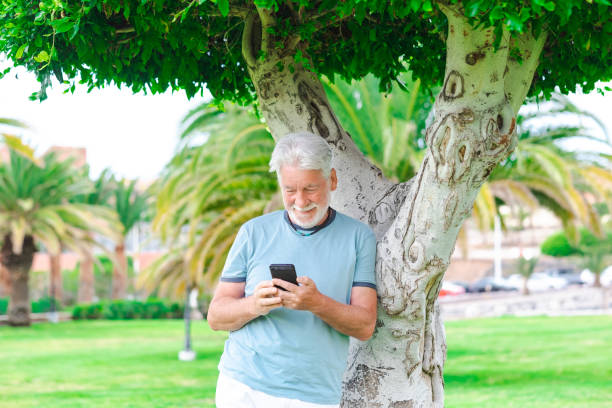istock Beautiful bearded senior man standing under a tree in public park using mobile phone. Smiling elderly man holding telephone using app and social media 1393755049