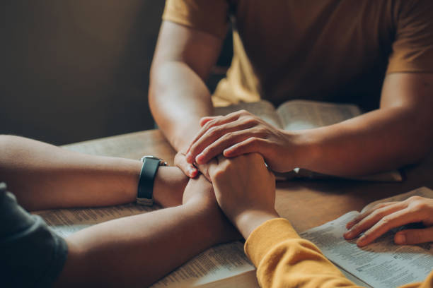 christian family sitting around a wooden table with open bible page and holding hands to bless and pray for each other. comforting and praying together.christians and bible study concept. - jesus christ human hand god consoling imagens e fotografias de stock