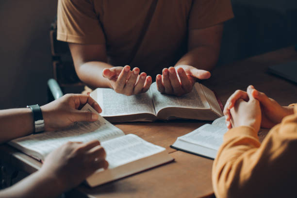 Christians and Bible study concept.Christian family sitting around a wooden table with open bible page and holding each other's hand praying together. Christians and Bible study concept.Christian family sitting around a wooden table with open bible page and holding each other's hand praying together. bible study group of people small group of people stock pictures, royalty-free photos & images