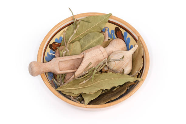 Garlic,  dried bay leaves, rosemary twigs and other spices, two empty small wooden spice spoons in the ceramic bowl on a white background