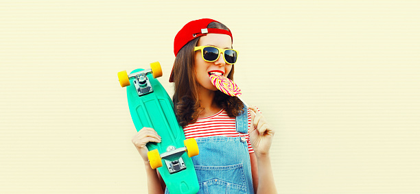Summer portrait of young woman with lollipop and green skateboard wearing baseball cap on white background