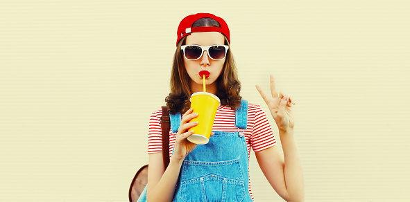 Portrait of young woman drinking fresh juice wearing red baseball cap and backpack on white background