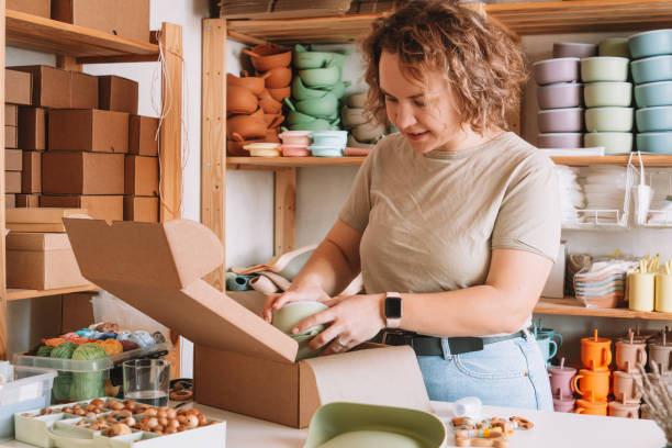 Smiling concentrated curly woman packing and wrapping shipment with silicone baby dish in box for delivery. Work at home stock photo