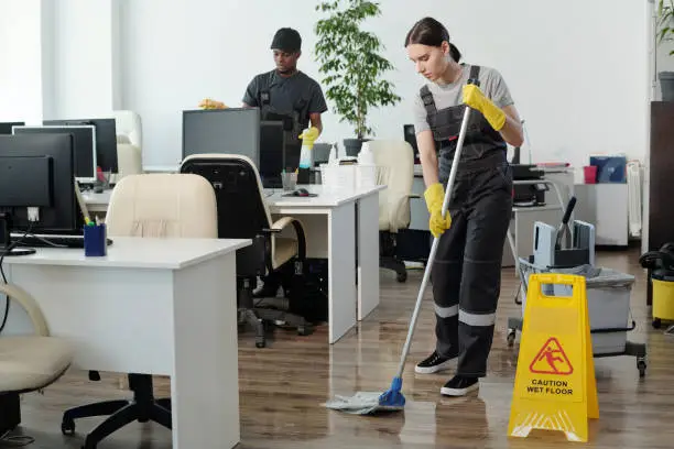 Photo of Young black man wiping computer monitors while woman with mop cleaning floor