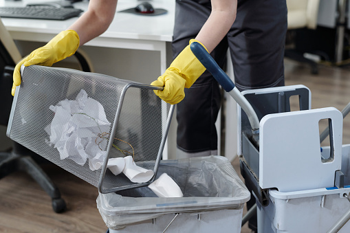 Gloved hands of female cleaner throwing trash from garbage bin into plastic bucket on janitor trolley while working in modern office