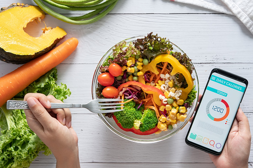 Calories counting , diet , food control and weight loss concept. Calorie counter application on smartphone screen at dining table with salad, fruit juice, bread and fresh vegetable. healthy eating