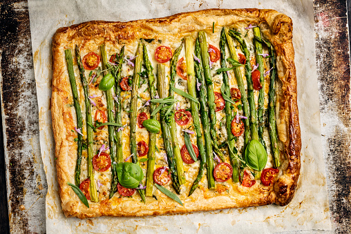 Overhead view of an asparagus, cheese and tomato tart. A shop bought puff pastry dough has been used as a base for the tart. Recipe; roll the  puff pastry out and make a thin cut not too deep about 2cm from the edge all around the pastry. Within the cuts prick the base of the pastry with a fork, lightly cover the pastry with some olive oil and herbs such as, chives, cress, thyme and parsley. Grate some cheese over the base such as gruyere or red cheddar and place the lightly cooked  asparagus spears that have have had the last few centimetres of woody stalk removed nose to tail on top of the cheese. Then chopped cherry tomatoes added, some shavings of fresh parmesan and some salt and pepper for seasoning. Fold over the 2 cm edges and brush some egg yolk over the pastry if there is any egg yolk left it can be poured over the asparagus. Cook for around 15 to 20 minutes at 180 degrees, delicious!