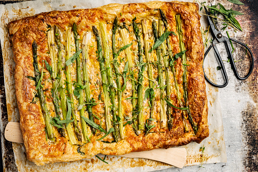 Overhead view of an asparagus and cheese galette. A shop bought puff pastry dough has been used as a base for the tart. Recipe; roll the  puff pastry out and make a thin cut not too deep about 2cm from the edge all around the pastry. Within the cuts prick the base of the pastry with a fork, lightly cover the pastry with some olive oil and herbs such as, chives, cress, thyme and parsley. Grate some cheese over the base such as gruyere or red cheddar and place the lightly cooked  asparagus spears that have have had the last few centimetres of woody stalk removed nose to tail on top of the cheese. Then add some shavings of fresh parmesan, salt and pepper for seasoning. Fold over the 2 cm edges and brush some egg yolk over the pastry if there is any egg yolk left it can be poured over the asparagus. Cook for around 15 to 20 minutes at 180 degrees, delicious!