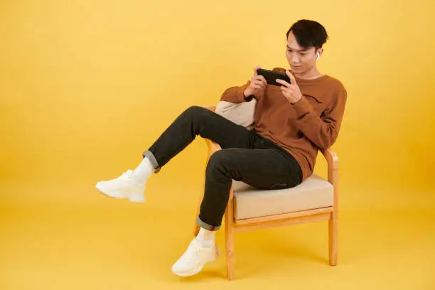 Vietnamese young man sitting on armchair and watching new show or playing game on smartphone