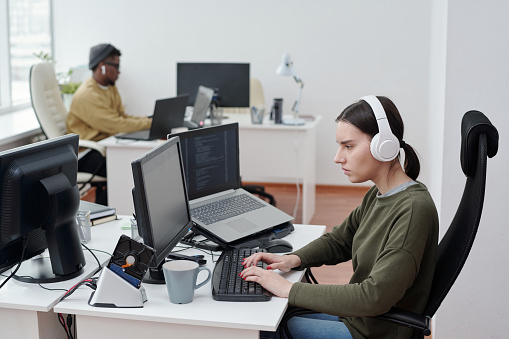 Side view of young female programmer in headphones listening to music by workplace against African American male colleague in office