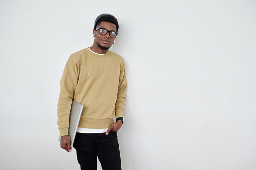 Young contemporary programmer or student in casualwear holding laptop while standing by wall of office and looking at camera